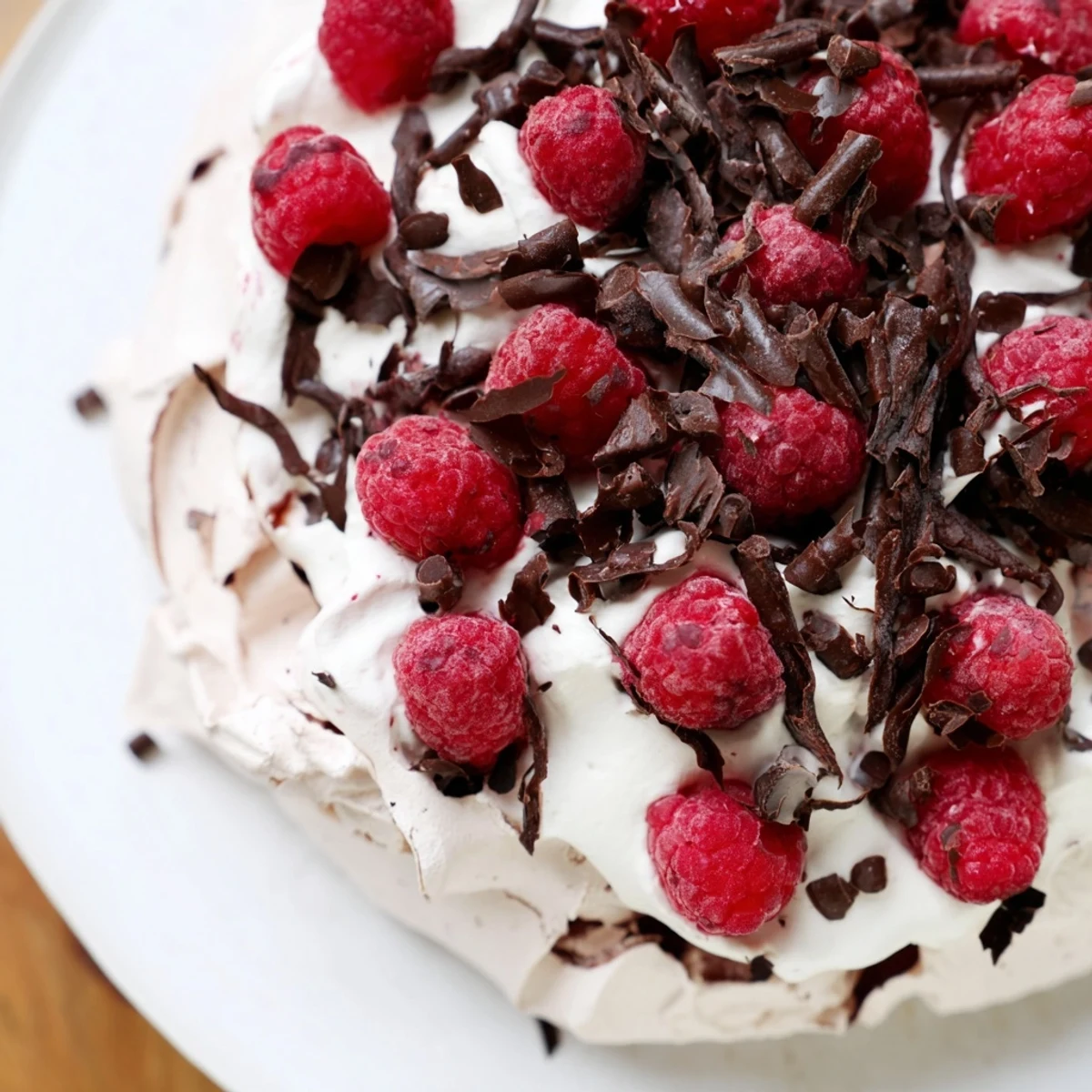 A slice of chocolate raspberry pavlova shows a crisp meringue shell piled high with cloud-like whipped cream, fresh red raspberries, and dark chocolate shavings, served on a white plate for a light dessert.