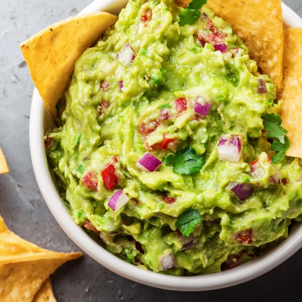 Bright, homemade tortilla chips surround a bowl of fresh Big Game Guacamole, loaded with ripe avocado, red onion, and cilantro.