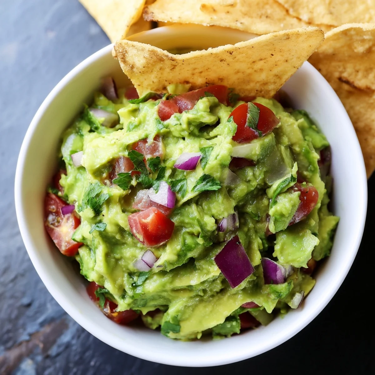 A top-down view of Big Game Guacamole garnished with lime wedges and a scattered pile of crispy, golden-brown corn chips.
