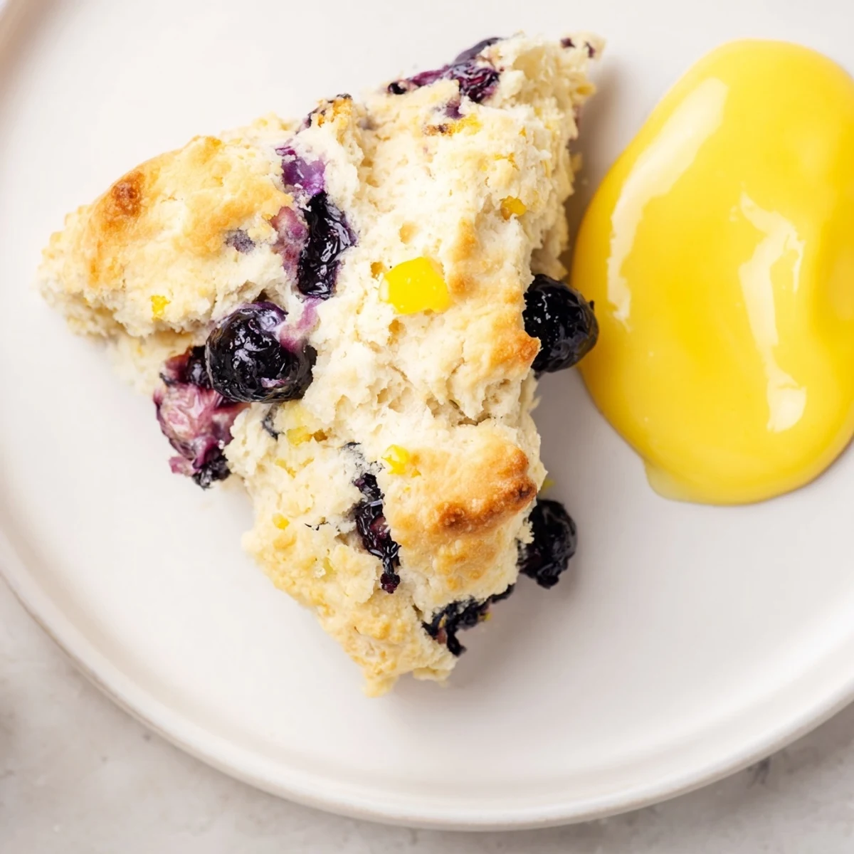 Buttery Lemon Blueberry Scones with Lemon Curd sit on a white plate, the crumb tender with visible blueberries and lemon zest flecks.
