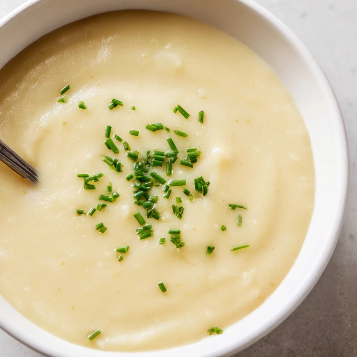 A rustic wooden table holds a ladle of Creamy Potato Soup with Chives, beside a glass of white wine.
