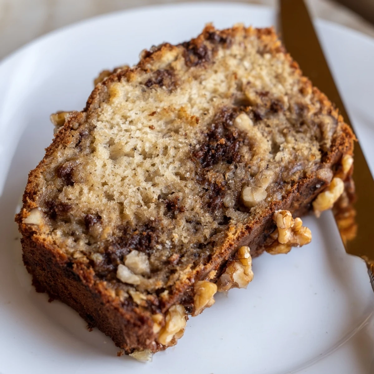 Thick slice of moist banana bread showing tender crumb, served warm on a white plate with optional walnuts