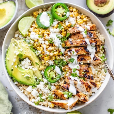 Close-up of Street Corn Chicken Bowl garnished with cotija cheese, cilantro, avocado slices, and lime wedges on a rustic table.